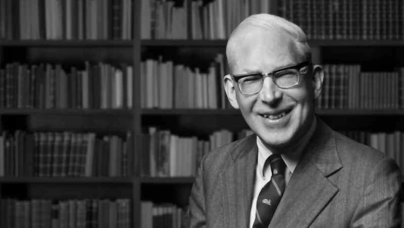 Fotografía en blanco y negro de J. I. Packer, sonriente, con gafas, traje y corbata, ante unas estanterías llenas de libros
